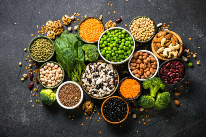 An assortment of legumes, nuts, and fresh vegetables arranged in bowls on a dark surface