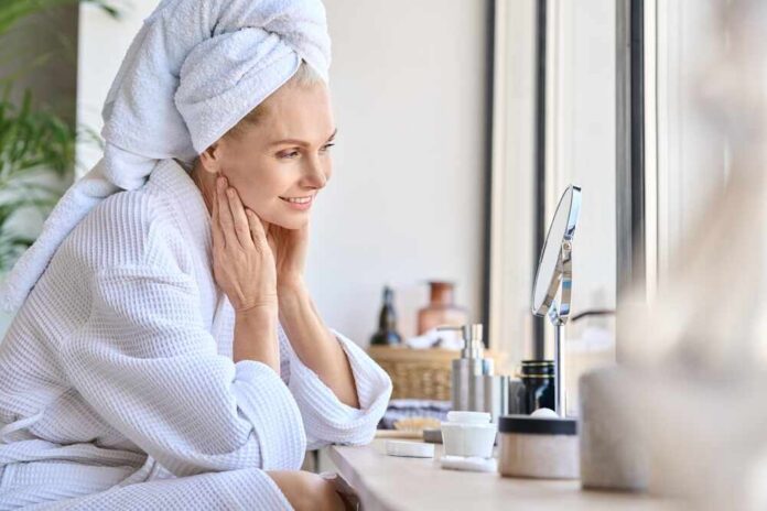shutterstock_2135514545.jpg Woman in a bathrobe with a towel on her head, smiling at her reflection in a mirror