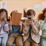 Four children sitting together, each focused on their electronic devices