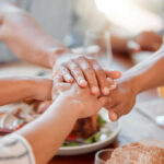 Several hands stacked together over a plate of food, symbolizing unity and support