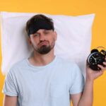 The Hidden Cost of Irregular Bedtime Patterns Man wearing a sleep mask holding an alarm clock with a frustrated expression