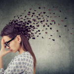 Silent Brain Fog Warnings Most Ignore A woman with glasses holding her head, with fragments dispersing from her head