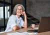 Gray Hair Rebellion Goes Mainstream Smiling woman with glasses at a desk with a laptop