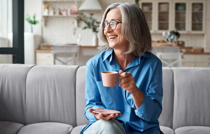 shutterstock_1818491870-1 Smiling older woman with gray hair holding a pink coffee cup on a sofa
