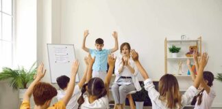 Children raise hands in a classroom circle with teacher clapping