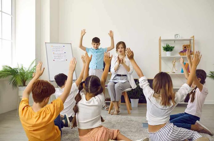 Children raise hands in a classroom circle with teacher clapping